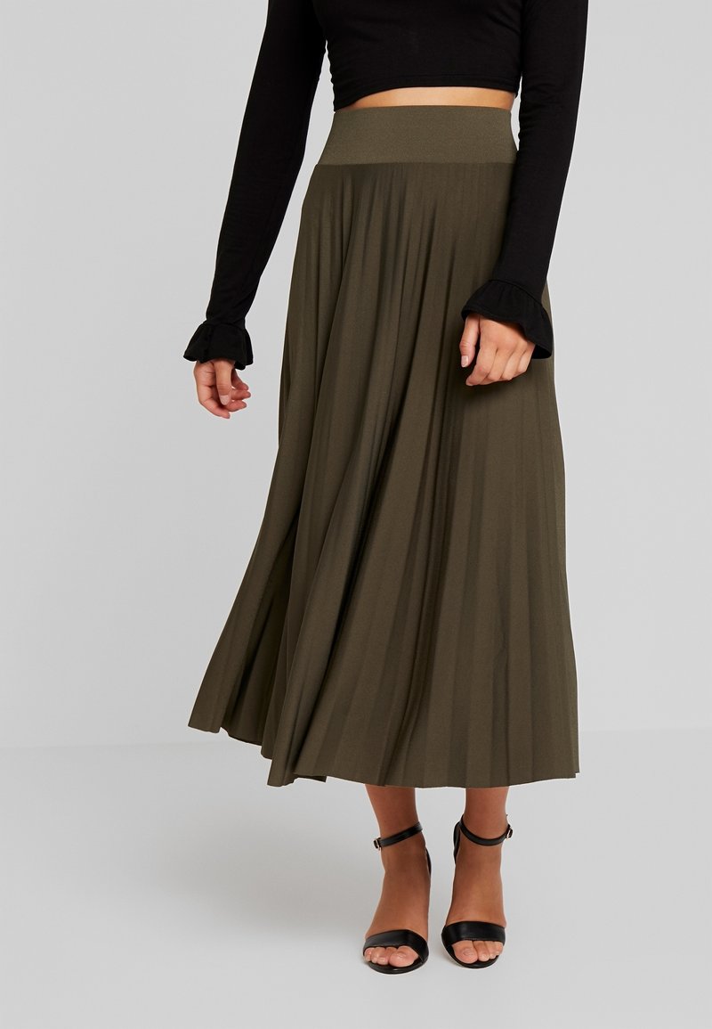 Femme portant une longue jupe plissée vert olive, un haut court à manches longues noires avec des poignets à volants, et des sandales noires à bride cheville.