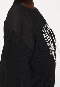 Sweat-shirt noir avec un design en tricot texturé à l'avant, des manches lisses contrastantes et un détail graphique blanc.