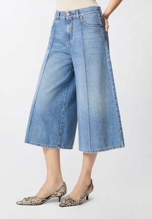 Γυναίκα που φοράει ανοιχτό μπλε τζιν παντελόνι με φαρδιά πόδια και cropped, μαζί με μυτερές, χαμηλοτάκουνες γόβες με σχέδιο φίδι, στέκεται σε λευκό φόντο.