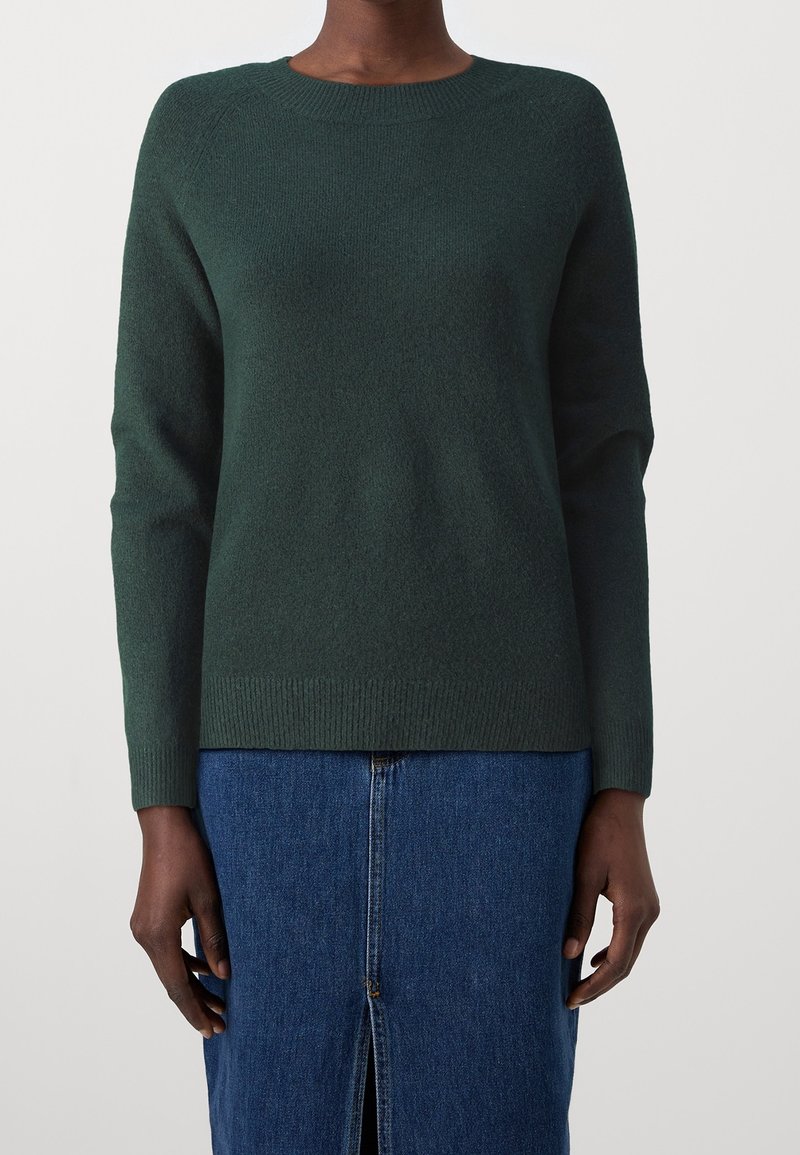 Personne portant un pull vert foncé à col rond et une jupe en jean bleue avec une fente à l'avant, debout devant un fond clair uni.