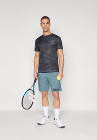 Camiseta deportiva negra con diseño, pantalones cortos teal, sosteniendo una raqueta de tenis y una pelota de tenis amarilla. Los zapatos son blancos con detalles oscuros.