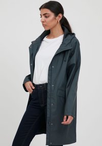 Waterdichte donkergroene regenjas met een capuchon, drukknoopsluiting en zijzakken. G gedragen over een wit T-shirt en donkere spijkerbroeken.