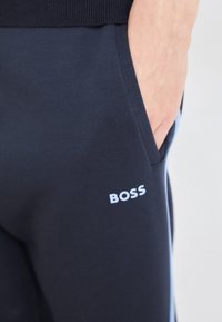 Tumši zilas sporta bikses, kas izgatavotas no mīkstas auduma, ar redzamu sānu kabatu un baltu "BOSS" logotipu uz augšstilba.