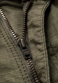 Green fabric with a textured pattern features a black YKK zipper, revealing interlocking teeth and a rectangular pull tab.