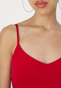 Un haut rouge à bretelles spaghetti avec un décolleté en V, une texture lisse et un minimum de détails ; associé à de grandes boucles d'oreilles dorées en forme de créole.