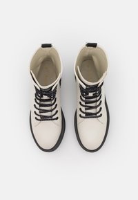 Botines de cuero blanco con cordones trenzados negros, punta redonda y suelas de goma negras. Cuenta con una lengüeta trasera para facilitar el calzado.