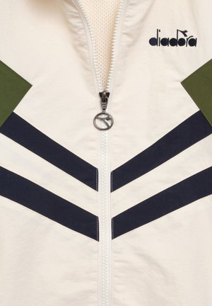 Giacca bianca con strisce a chevron nere, dettagli verdi sulle spalle, una cerniera con tiretto circolare argentato e il logo "diadora" nero sul petto.