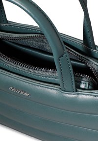 Borsetta in pelle teal con doppi manici, dotata di un comparto con zip, cuciture testurizzate e un piccolo logo argento sul lato.