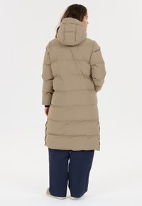 Lang beige puffjakke med hætte, der har et quiltet design. Båret over mørkeblå brede bukser med sideslids i kanten.