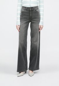 Sive široke kavbojke z izbledelim zaključkom, z gumbno zaporo in dizajnom s petimi žepi. Združene s svetlimi čevlji s koničastim prstom.