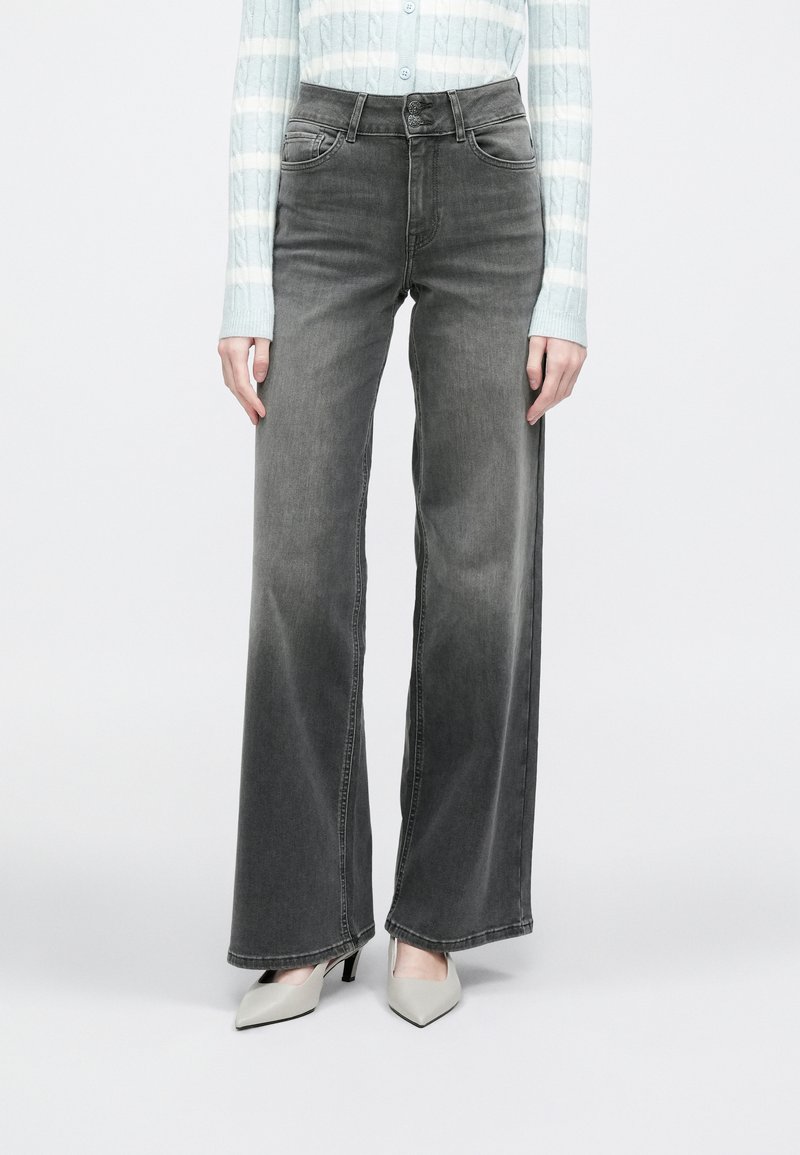 Sive široke kavbojke z izbledelim zaključkom, z gumbno zaporo in dizajnom s petimi žepi. Združene s svetlimi čevlji s koničastim prstom.