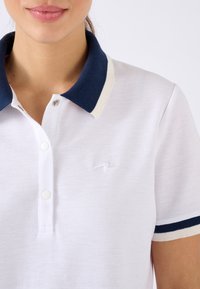 Polo blanc en tissu texturé avec un col bleu marine. Présente une bordure crème et un petit logo brodé sur la poitrine.