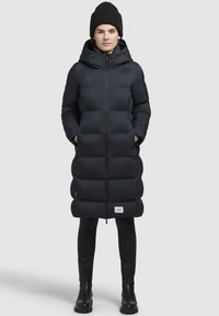 Dlhý, tmavomodrý pufový kabát s kapucňou, so zipsom vpredu, prešitým dizajnom a bočnými vreckami. Nosený nad čiernymi legínami a čižmami.