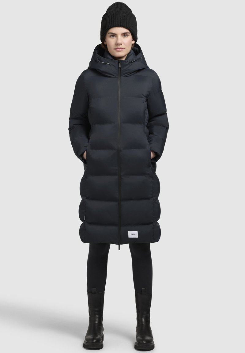 Dlhý, tmavomodrý pufový kabát s kapucňou, so zipsom vpredu, prešitým dizajnom a bočnými vreckami. Nosený nad čiernymi legínami a čižmami.