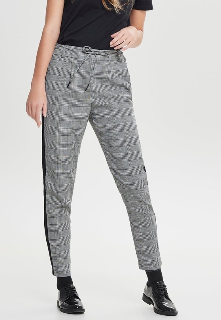 Femme portant un pantalon à carreaux gris avec des bandes latérales noires, des chaussettes noires, des chaussures noires brillantes à lacets, et un chemisier noir à manches courtes.