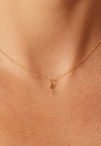 Collier pendentif en or avec un charme en forme de lettre "E", sur une fine chaîne, posé sur un fond couleur peau.