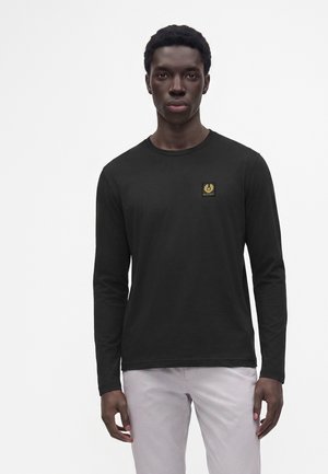 Junger Mann mit kurzen Haaren, der ein schwarzes Langarmshirt mit kleinem gelben Logo auf der Brust und helle Hosen vor weißem Hintergrund trägt.