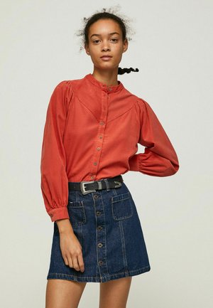 Blusa button-up color ruggine con maniche a sbuffo, abbinata a una gonna di jeans blu con bottoni frontali e tasche, accentuata da una cintura nera.