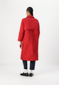 Czerwony płaszcz zapinany na guziki z nakładką na plecach, wykonany z lekkiej tkaniny, z długimi rękawami i rozcięciami po bokach, noszony z granatowymi spódnicowymi spodniami i czarnymi butami.