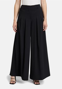 Zwarte wijde pantalon met voorplooien, tailored fit en een gladde textuur. Bevat riemlussen en een ritssluiting. G gedragen met slanke hakken.