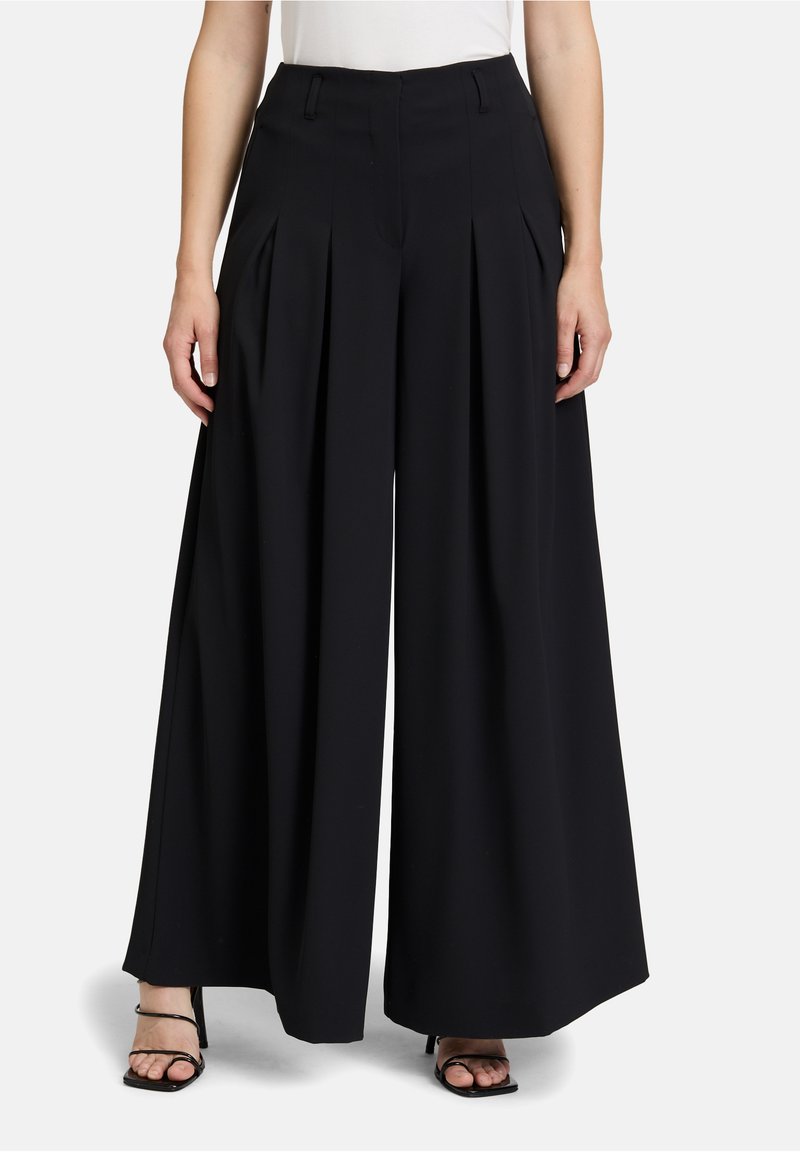 Zwarte wijde pantalon met voorplooien, tailored fit en een gladde textuur. Bevat riemlussen en een ritssluiting. G gedragen met slanke hakken.