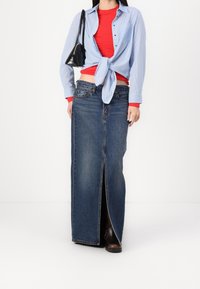 Jupe longue en jean avec une fente à l'avant, portée avec un top rouge ajusté et une chemise à rayures bleu pâle boutonnée et nouée à la taille. Sac à main noir et bottes.