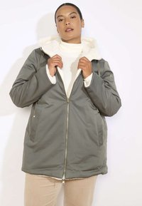 Olivgrüne, isolierte Jacke mit einem bequemen, weißen Fleece gefütterten Kapuze, seitlichen Taschen und einem goldenen Reißverschluss. Weiche, glatte Textur.
