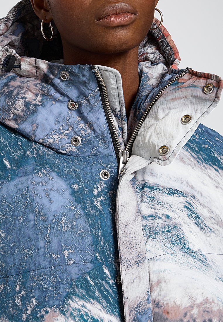 Gros plan sur une personne portant une veste d'hiver texturée bleue et blanche avec des fermetures éclair argentées, des boutons pression et des boucles d'oreilles visibles.