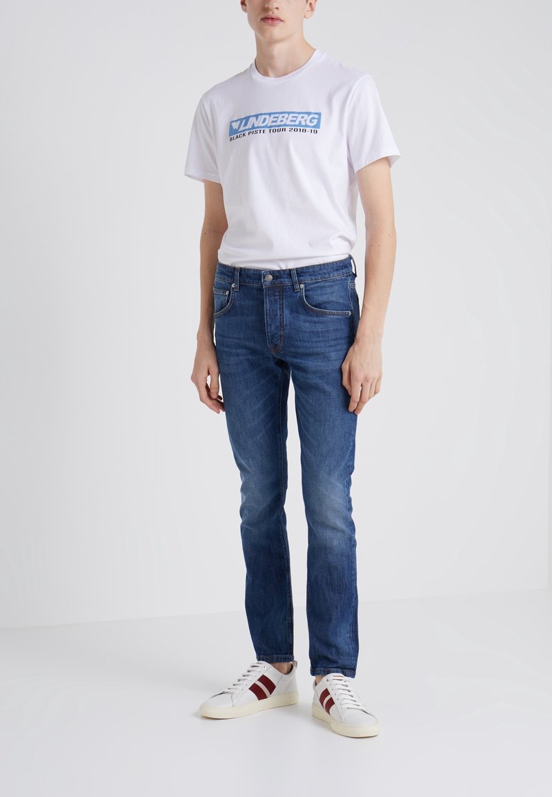 Vit t-shirt med blå logotext, kombinerad med mörkblå denimjeans. Bärs med vita sneakers med röda ränder. Tight passform.