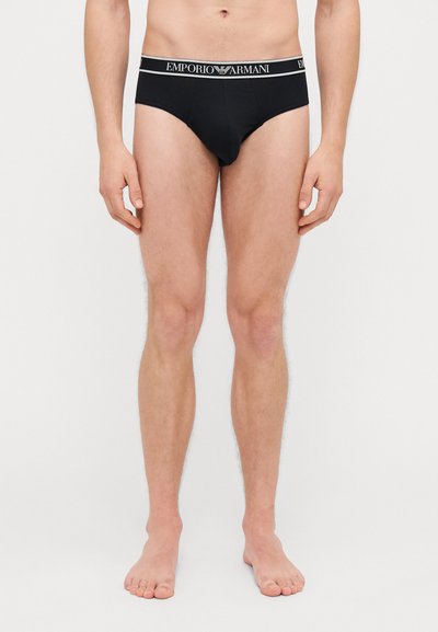 Slip in cotone nero con cinturino bianco con il marchio "EMPORIO ARMANI". Il design è aderente con una forma classica del slip e cuciture minime.