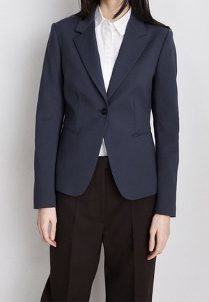Vrouw draagt een marineblauwe, nauwsluitende blazer over een wit overhemd met kraag en zwarte broek, staand tegen een effen achtergrond.
