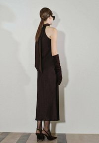 Vestido negro de tirantes con una lazada trasera fluida, combinado con guantes largos negros y zapatos de tacón alto tipo slingback, posicionado contra una pared lisa.