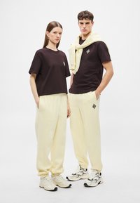 Δύο μοντέλα φορούν ταιριαστά σύνολα: κοντά μπλουζάκια σε σκούρο καφέ, κίτρινες παντελόνες με ελαστική μέση και χοντρά λευκά αθλητικά παπούτσια με μαύρες λεπτομέρειες.