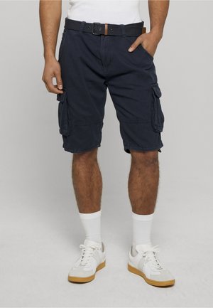 Mann trägt navyblaue Cargo-Shorts mit Klappentaschen, einen schwarzen Gürtel, weiße Socken und weiße Sneakers mit Gummisohlen, steht mit einer Hand in der Tasche.