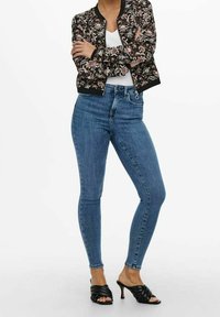 Veste bombardier florale avec une base noire, un dessous blanc, un jean skinny bleu et des sandales noires à talons ouverts, en pose angulaire.