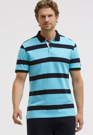 Mann trägt hellblaues Polohemd mit breiten, marineblauen horizontalen Streifen und dunkelblauem Kragen, steht vor einfachem hellem Hintergrund.