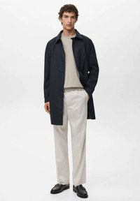 Cappotto blu scuro con chiusura a bottoni, maglione beige chiaro, pantaloni crema e scarpe nere. Design semplice e dritto, con dettagli minimi.