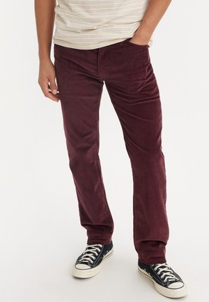 Osoba ubrana w ciemnoczerwone spodnie sztruksowe i czarne niskie trampki ze sznurowadłami w kolorze białym, jedna ręka w kieszeni, stojąca na białym tle.