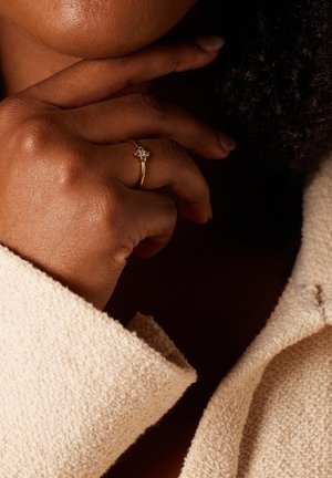 Gouden ring met een bloemmotief en drie heldere stenen, gedragen op een hand die de kin aanraakt, tegen een textuur van romige stof.