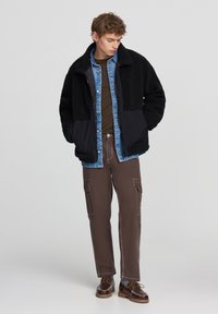 Giacca sherpa nera con una camicia di jeans blu navy sotto, pantaloni cargo marroni e scarpe stringate marroni. Le texture sono varie e sovrapposte.