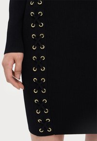 Black ribbed dress with gold grommets and crisscross black laces on the side. Close-up shows hand positioned near the design detail.