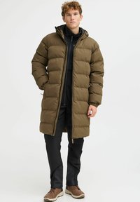 Long, hooded puffer coat in olive green, featuring quilted texture, front zipper, and elastic cuffs, paired with black pants and brown footwear.