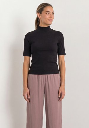 Frau steht mit entspannten Händen, trägt ein schwarzes kurzärmeliges Rollkragentop und lockere rosa weite Hosen, blickt zur Seite.