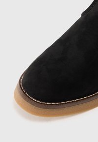 Chausson en suédine noire avec un bout arrondi, semelle en caoutchouc beige contrastante et couture beige visible le long du bord.