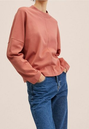 Osoba w luźnej różowej bluzce z długim rękawem i niebieskich dżinsach z wysokim stanem, z rękami w kieszeniach, na jednolitym tle.