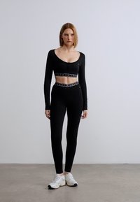 Μαύρο crop top με μακριά μανίκια και στρογγυλή λαιμόκοψη, συνδυασμένο με μαύρο κολάν εφαρμοστό. Και τα δύο έχουν ζώνη μέσης με αντιθετικό λογότυπο. Το μοντέλο φοράει άσπρα αθλητικά παπούτσια.
