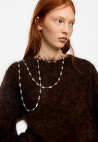 Mujer joven con cabello rojo que lleva un suéter marrón oscuro y peludo y un collar largo de plata con cuentas claras colgado sobre el pecho y el cuello.