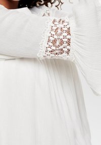 Blouse blanche avec des manches longues évasées ornées d'une dentelle au poignet, en tissu texturé et avec des plis sur le corps pour un aspect fluide.