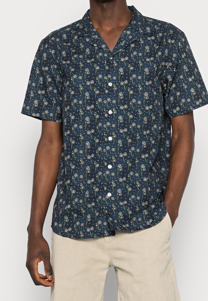 Homme portant une chemise bleu marine à manches courtes avec un petit motif floral et un pantalon beige clair.