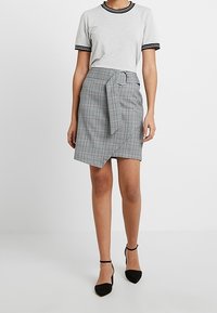 Grijze T-shirt met zwarte streepjessluiting, gecombineerd met een geruit wikkelrok met een riem en een ringdetail. Zwarte puntige flats.